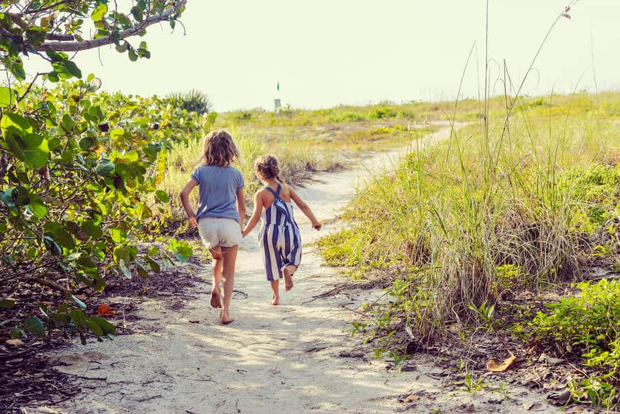 Two girls walk hand in hand down a sandy path, surrounded by nature.